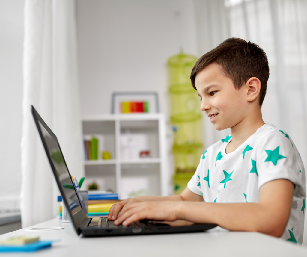 A boy between 8-10 wearing a white T-shirt with green stars on it smiles while typing on his laptop. A shelf of books and blocks is in the background.